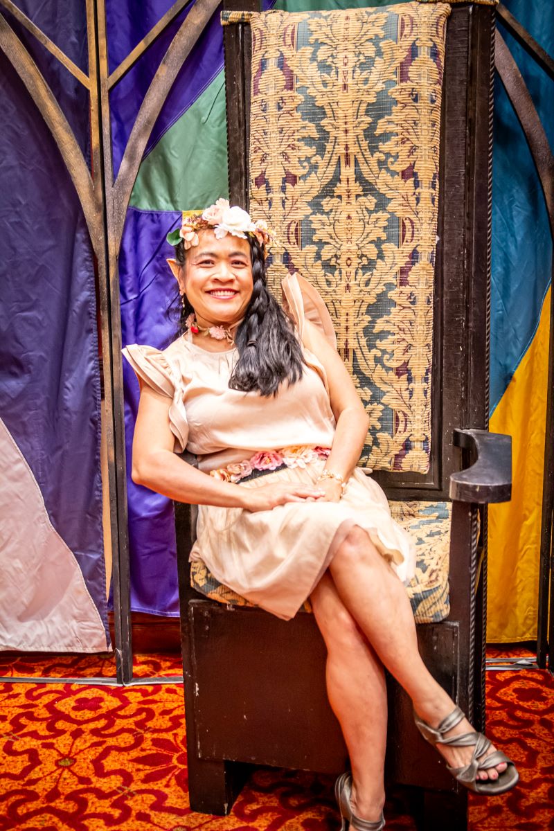 A woman dressed as an elf with a flower crown and pink dress sits in a throne chair in the club area of Soonercon.
